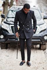Stylish african american gentleman in elegant black jacket, holding retro walking stick as cane flask with golden diamond ball handle. Rich fashionable afro man against business suv car.