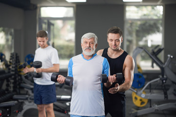 Fototapeta na wymiar Trainer helping to work out elderly man with dumbbells.