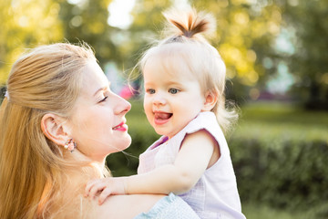 Young beautiful blonde mother with her baby girl laughing together and playing in green park outdoors at sunny day