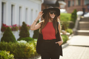 Beautiful brunette model girl with perfect body in the red blouse and in the fashionable black suit adjusting her stylish black hat, smiles, and posing at sunny city street