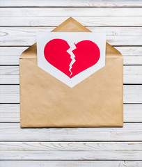 Big brown envelope with a sheet of paper which shows a broken red heart. Vintage white wooden background. The concept of breaking relationships, betrayal, divorce, separation and nostalgia.