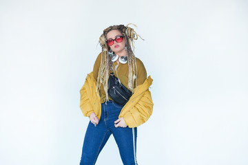 young blonde girl in sunglasses with african pigtails listens to music in headphones and dances against a white background.