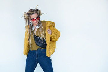 young blonde girl in sunglasses with african pigtails listens to music in headphones and dances against a white background.