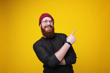 Smiling bearded hipster man with round glasses pointing away at copyspace over yellow background
