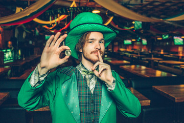 Happy rich young man in St. Patrick's suit stand in pub and hold golden coin. He has finger close to mouth. Guy look on camera. He stand alone.