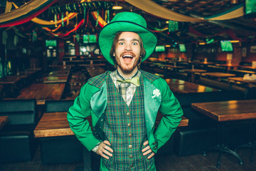 Happy excited young man hold hands on hips and smile to camera. He wear St. Patrick's suit. Guy stand alone in pub.