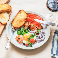 Fresh rustic breakfast - fried egg with bacon, onion and tomatoes on a plate, a glass of red wine and toast on a light wooden background