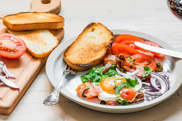 Fresh rustic breakfast - fried egg with bacon, onion and tomatoes on a plate, a glass of red wine and toast on a light wooden background