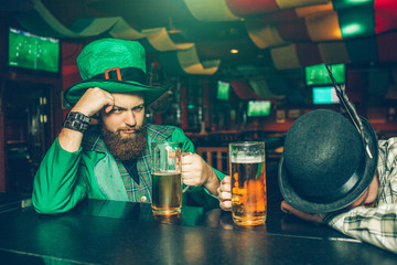 Drunk young man in green suit sit at bar counter in pub with friend. Another guy fell asleep. They have mugs of beer.