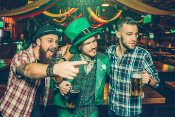 Excited young men stand in pub together. Guy on left point. They look to right. Young man in green suit wear St. Patrick's costume.