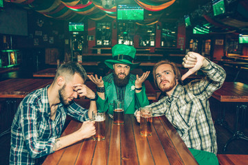 Upset and unhappy young men sit at table in pub. They have mugs of beer. Man in middle wear St. Patrick's suit.