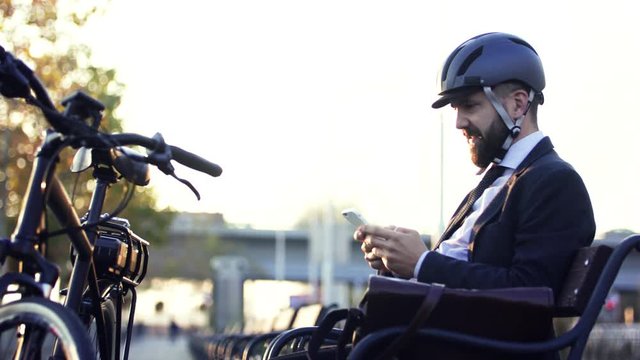Businessman commuter with bicycle helmet sitting on bench in city, using smartphone.