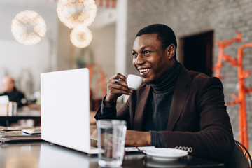 Portrait of happy african businessman using phone and drinking coffee while working on laptop in a restaurant.
