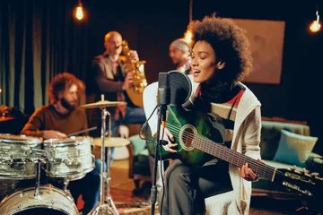 Fotobehang Mixed race woman singing and playing guitar while sitting on chair with legs crossed. In background drummer, saxophonist and bass guitarist. © chika_milan
