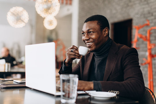 Portrait of happy african businessman using phone and drinking coffee while working on laptop in a restaurant.