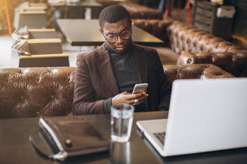Portrait of happy african businessman using phone while working on laptop in a restaurant.