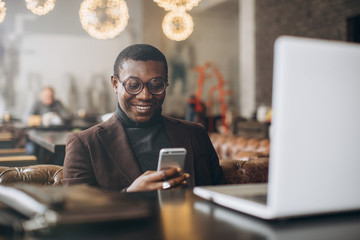 Portrait of happy african businessman using phone while working on laptop in a restaurant.