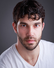 Obraz premium Headshot of handsome caucasian man with stubble and black hair and white t shirton grey background