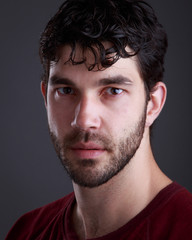 Headshot of handsome caucasian man with stubble and black hair and burgundy t shirt on grey background