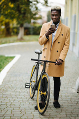 Naklejka premium Portrait of handsome young african man using mobile phone and fixed gear bicycle in the street.