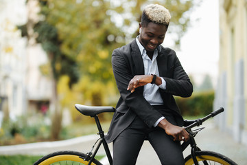 Fototapeta premium Portrait of young handsome african man with bicycle standing outdoors looking at watch.