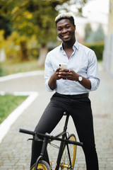 Handsome young Afro American man in casual clothes is using smartphone, looking at camera and smiling while leaning on his bike, standing outdoors