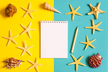 Notebook and pencil with seashells and sea stars on blue and yellow paper background. Mock up, flat lay, copy space, top view. Summer vacation concept