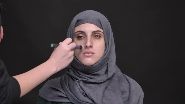 Portrait of female hands doing make-up for middle-aged muslim woman in hijab on black background.