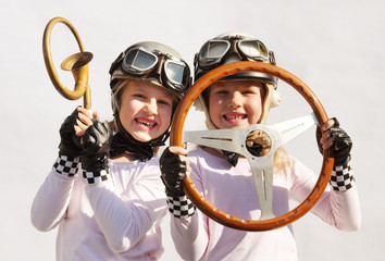 Twin sisters play race car driver sitting in their imaginative race car. They play happily wearing vintage helmets, goggles and use an old-timer steering wheel they found in the garage.