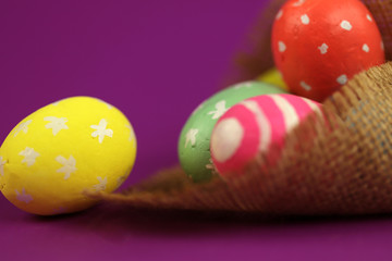 Easter background. Green, red and yellow painted eggs handmade in burlap on purple background with bokeh. Cropped shot, close-up, nobody, horizontal, blurred, free space. Easter concept.