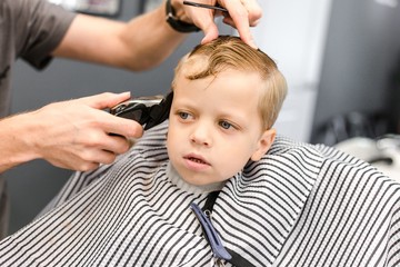 Obraz na płótnie Canvas Hairdresser scissors cuts the hair of a little boy blonde with blue eyes making a fashionable hairstyle in the salon of male beauty