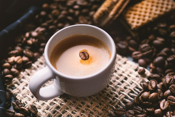 
    Cup of coffee in the middle of coffee beans with biscuits and tablecloth. Grained product. Hot drink. Close up. Harvesting. Natural background. Energy. Fresh espresso with beans on the foam.