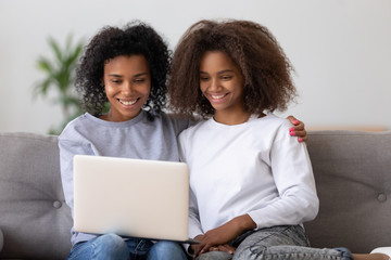Fototapeta na wymiar Smiling African American mother and daughter sit on couch watching video on laptop, black millennial nanny hang out with teen girl, relax on sofa using computer, mom and kid rest together at home