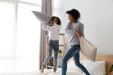 Excited African American mom and daughter have pillow fight jumping on bed, happy black nanny and...