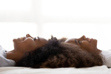 Happy African American mom and daughter lying on their backs on bed touching heads, smiling young mommy and teen girl relax at home together, nanny or friend spend time with teen kid dreaming