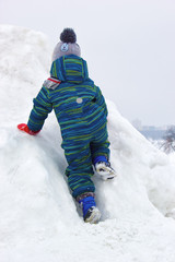 Fototapeta na wymiar A four-year-old child, a boy, is climbing a snowy hill. He is wearing colorful winter clothes. View from the back