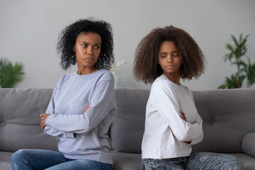 Angry African American mother and daughter sit back to back on couch, avoid talking or looking, mad...