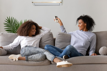 Smiling African American mom and daughter sit on cozy couch relaxing, operating air conditioner...