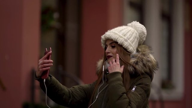Adorable girl with braces in knitted hat and parka is standing at the streer and talking on a video chat and smiling. Girl is holding her phone in front of her and wearing an earphones.
