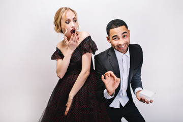 Portrait handsome man in tuxedo with gift smiling to camera behind astonished young woman in luxury dress on white background. Valentine s day, love, dating, having fun, surprise, present