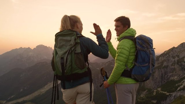 SLOW MOTION, CLOSE UP, LENS FLARE: Young couple hiking in the stunning mountains high five after reaching the mountaintop at sunset. Cheerful trekkers celebrate seeing the sunrise during their ascent.
