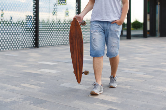 Cropped outdoor picture of man in denim shorts and grey sneakers walking with a longboard in right hand/ urban area, moving fast, hipster skateboarding in city/ people, lifestyle and sport concept.