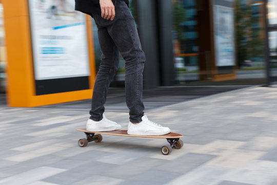 Shot of male in black jeans and white sneakers riding a longboard in urban area, photo in motion/ side view/ wooden longboard, moving fast, city center, enjoy cruising, people and sport concept.