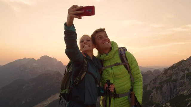 SLOW MOTION, LENS FLARE, PORTRAIT: Goofy hiker couple making funny faces while taking selfies on the summit at sunset. Happy man and woman taking funny pictures on a sunny morning in the mountains.