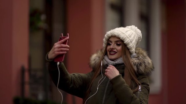 Cute girl is holding her phone in front of her and wearing an earphones. Girl is wearing braces, knitted hat and parka is standing at the streer and talking on a video chat and smiling.