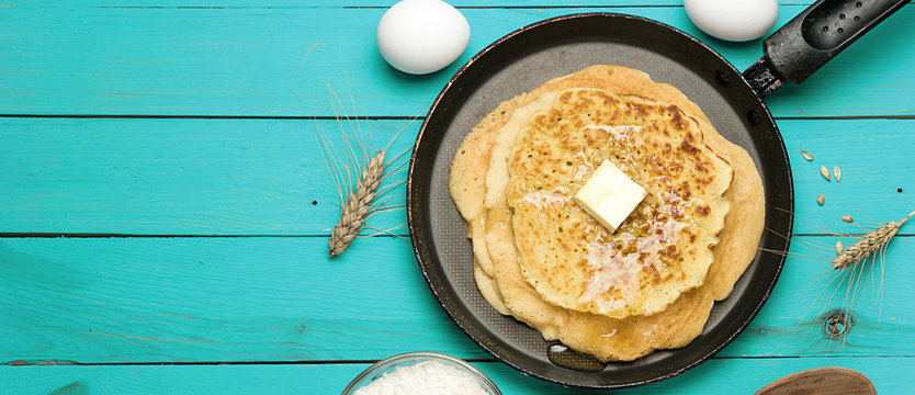 Hot delicious pancakes in frying pan on blue wooden table with flour and eggs, panoramic banner.