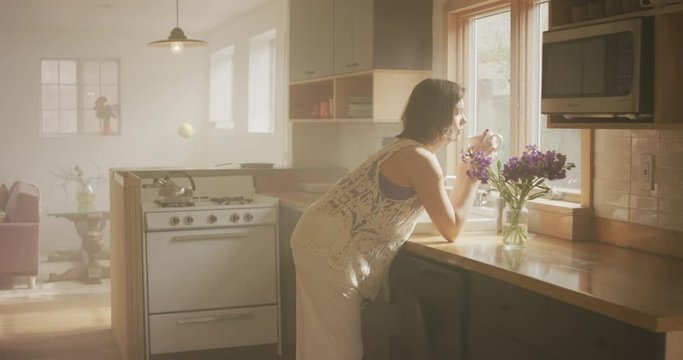A woman leans on her counter in her modern kitchen.  She is looking out the window.  She turns and looks at a vase of purple flowers on her counter.