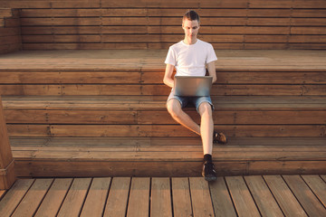 Portrait of a happy young male freelancer working on the street near the sea on a wooden platform with a laptop and smartphone on a sunny summer day. Remote work concept