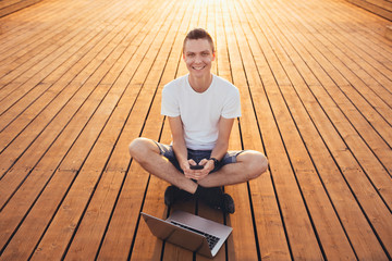 Portrait of a happy young male freelancer working on the street near the sea on a wooden platform with a laptop and smartphone on a sunny summer day. Remote work concept