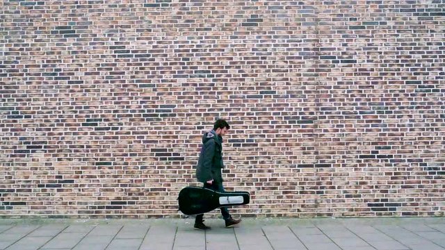 Musiker, Gitarrist geht mit seiner Gitarre an einer Backsteinmauer entlang